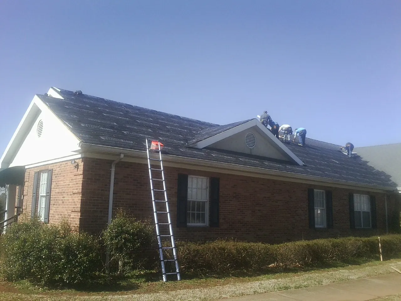 Crew tearing shingles off a brick ranch roof with a ladder set — Metal Roof Repair work by our crew in West Rutland
